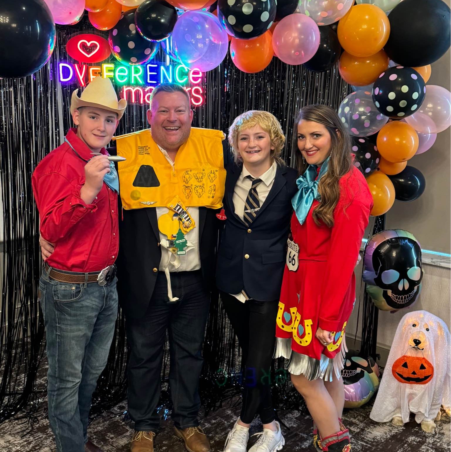 Four individuals in playful costumes stand before a vibrant Halloween-themed backdrop at a Dyfference Makers community event. Surrounded by balloons, fringe curtains, and a neon sign reading “LOVE DIFFERENCE matters,” they embody the nonprofit’s mission to celebrate neurodiversity and support those with dyslexia through joyful, inclusive gatherings.